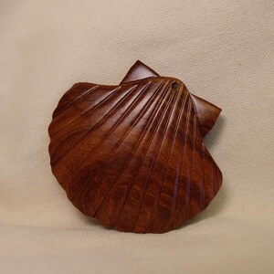 May include: A handcrafted, wooden seashell. The brown, carved scallop shell has detailed ridges and a natural finish. The shell is a decorative item, possibly for coastal or nautical themed decor.
