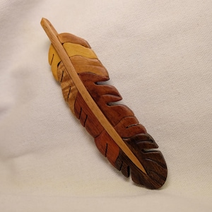 May include: A wooden feather with a natural color gradient, from light tan to dark brown. The feather is intricately carved with detailed edges and a central spine. The item is displayed on a white fabric background.