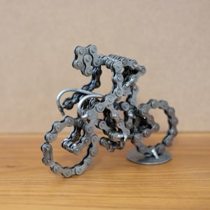 May include: A metal sculpture of a cyclist riding a bicycle, crafted from bicycle chains. The sculpture features a detailed representation of a cyclist on a bike, with the chain links forming the body and wheels. The sculpture is on a wooden surface.