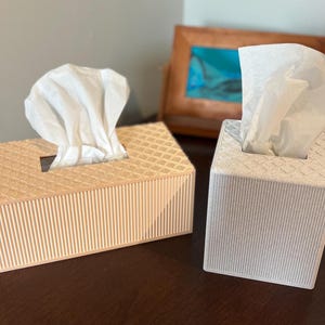 May include: Two decorative tissue box covers. One is light beige with a textured top and vertical ribbed sides. The other is white with a similar textured top and ribbed sides. Both have white tissues protruding from the top opening.