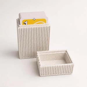 May include: A rectangular, off-white, speckled storage box with vertical ribbed sides. The box is open, with a matching lid placed beside it. A stack of playing cards with a yellow design and the number one is visible inside the main container.