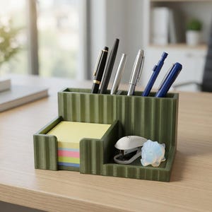 May include: A green desk organizer with multiple compartments. It holds pens, a stack of sticky notes, a stapler, and a decorative stone. The organizer is made of a ridged material and sits on a light-colored wooden desk.