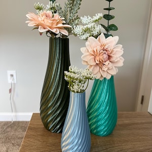 May include: Three decorative vases in shades of green and blue, each with a spiral design. The vases hold faux flowers in peach and white. The vases are arranged on a wooden surface.