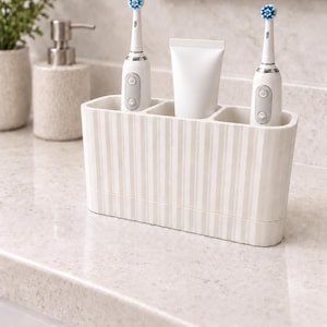 May include: A rectangular bathroom organizer with a vertical striped pattern in off-white and beige. The organizer features three distinct compartments holding two white electric toothbrushes with blue bristles and one plain white toothpaste tube.