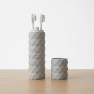 May include: A gray, geometric-patterned toothbrush holder with two white toothbrushes and a matching cup. The holder is cylindrical and has a textured surface. The cup is small and also has a geometric design. The items are on a light brown surface.