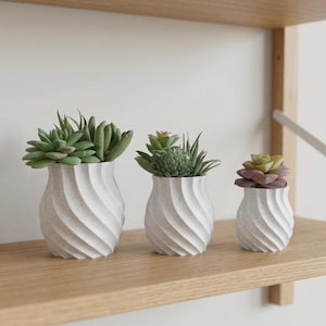 May include: A set of three ceramic mugs with a speckled, matte finish. The mugs are in shades of gray, white, and black. Each mug has a simple, minimalist design, perfect for coffee or tea. The mugs are stacked on top of each other.