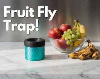May include: A teal cylindrical fruit fly trap with a black lid and a textured honeycomb pattern on the exterior. The text "Fruit Fly Trap!" is displayed in white letters above the trap, which sits near a glass bowl of apples, grapes, and bananas.