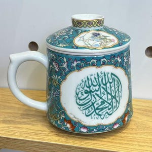 May include: A ceramic tea mug with a matching lid, featuring a teal and floral design. The mug has a white handle and a central white oval with green Arabic calligraphy. The mug sits on a wooden surface.