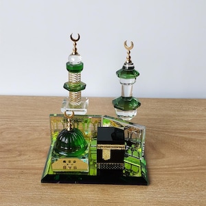 May include: A crystal mosque model with green and gold accents. The model features two minarets, a dome, and a small cube representing the Kaaba. The base is decorated with a green and gold pattern.