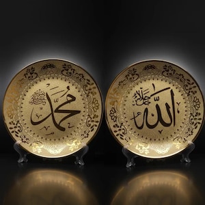 May include: Two decorative gold-colored plates with Arabic calligraphy. One plate features the name "Muhammad", and the other displays the word "Allah". Both plates have ornate floral patterns around the edges and are displayed on clear stands.