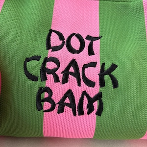 May include: A knitted fabric featuring vertical stripes in alternating shades of bright green and light pink. Centered on the fabric is black embroidered text that reads DOT CRACK BAM in a bold, slightly irregular, hand-drawn style font.