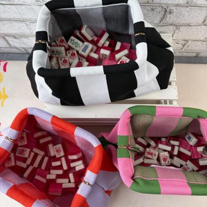 May include: Three fabric pouches with zippered openings, each filled with small rectangular mahjong tiles. One pouch features black and white vertical stripes, another has orange and light purple blocks, and the third has green and pink blocks.
