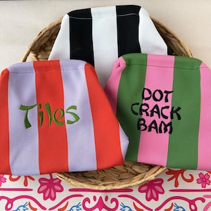 May include: Three fabric pouches with vertical stripes. One pouch has black and white stripes. Another has red and light purple stripes with the green embroidered text Tiles. The third has pink and green stripes with the black embroidered text DOT CRACK BAM.