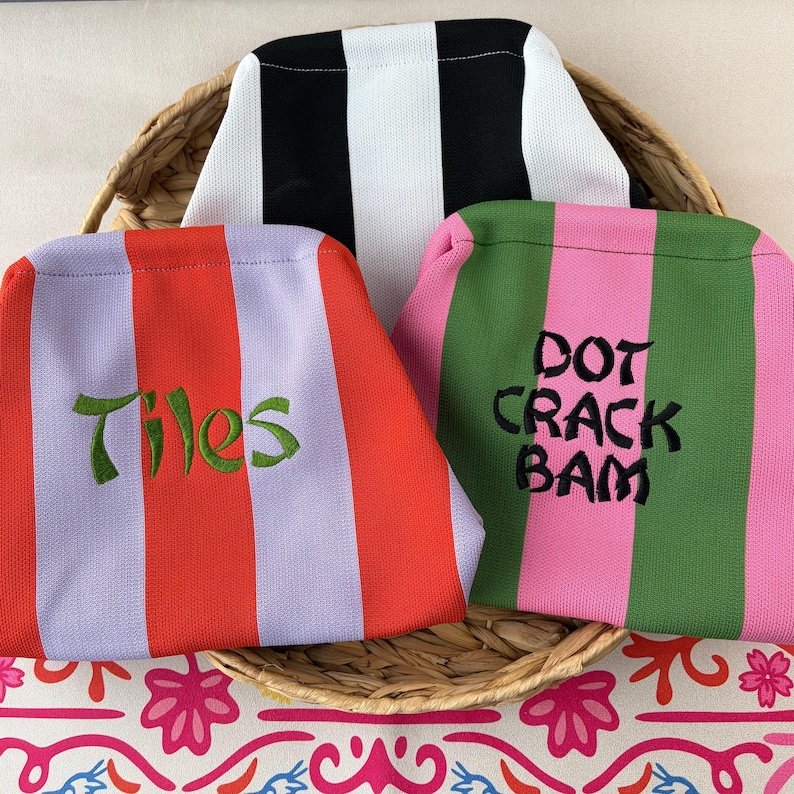 May include: Three fabric pouches with vertical stripes. One pouch has black and white stripes. Another has red and light purple stripes with the green embroidered text Tiles. The third has pink and green stripes with the black embroidered text DOT CRACK BAM.