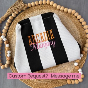 May include: A black and white striped fabric pouch featuring the embroidered text ARCADIA in orange and Mahjong in pink. The pouch has a structured shape and is centered on a circular woven mat surrounded by a decorative wooden bead garland.