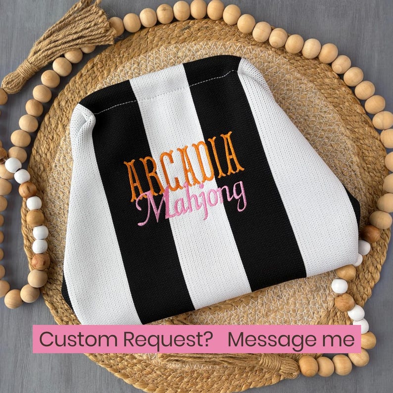 May include: A black and white striped fabric pouch featuring the embroidered text ARCADIA in orange and Mahjong in pink. The pouch has a structured shape and is centered on a circular woven mat surrounded by a decorative wooden bead garland.