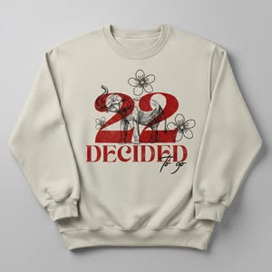 Op de afbeelding: Crèmekleurige sweatshirt met een rode afbeelding. Het ontwerp bevat het nummer "22", een olifantenillustratie en de tekst "DECIDED to go". Bloemenaccenten zijn ook in het ontwerp opgenomen.