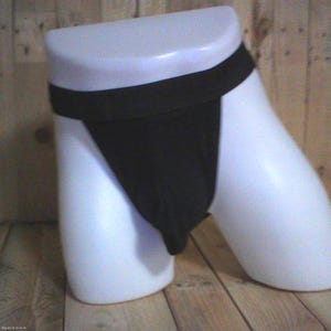 Op de afbeelding: Een effen zwarte jockstrap met een brede, platte tailleband en een enkele voorzak. Het kledingstuk wordt getoond op een witte, gladde, torso-vormige etalagepop, wat het eenvoudige ontwerp en de diepzwarte stoffen constructie benadrukt.
