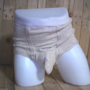 May include: A pair of beige ribbed underwear featuring a high-waisted design with a thick elastic waistband and vertical stitched detailing along the front, displayed on a white mannequin torso against a light-coloured wooden background.
