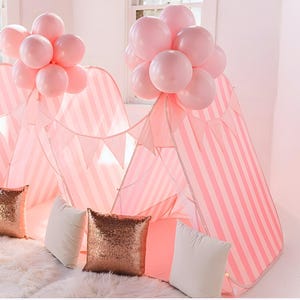 Puede incluir: Una tienda de campaña de juego a rayas rosas y blancas, adornada con globos rosas y cojines decorativos. La tienda tiene forma de tipi, con una serie de rayas rosas y blancas. Los globos son de color rosa suave y los cojines son blancos y con lentejuelas.