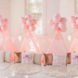 Puede incluir: Una escena interior caprichosa con tiendas de campaña tipi rosas, adornadas con globos rosas y cojines decorativos. Los cojines presentan diseños de lazo y ballet, y detalles con lentejuelas. La escena está ambientada sobre una alfombra blanca y esponjosa.