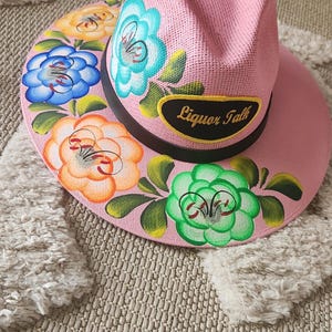 May include: Pink straw hat with a black band and a yellow patch that says "Liquor Talk". The hat is decorated with hand-painted flowers in shades of blue, green, orange, and yellow.