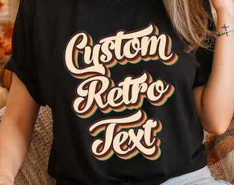 May include: A black short-sleeved t-shirt featuring the words Custom Retro Text printed in a layered, multi-colored font with cream, orange, and teal outlines. The shirt has a crew neckline and is tied in a knot at the bottom hem.