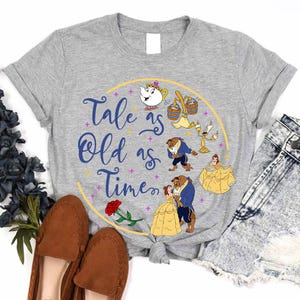 Op de afbeelding: Grijs t-shirt met een cirkelvormig ontwerp met personages uit Belle en het Beest. Het ontwerp bevat de tekst "Tale as Old as Time" in blauw script, en illustraties van de personages in geel en blauw.