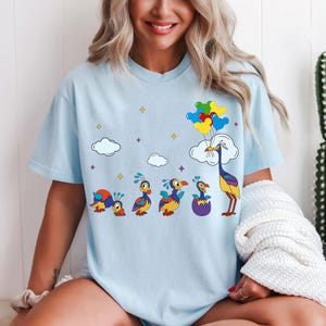 Pode incluir: Camiseta azul claro com uma ilustração de desenho animado do ciclo de vida de um pássaro. O design inclui uma série de pássaros em vários estágios, desde uma forma semelhante a uma lagarta até um pássaro segurando balões. Nuvens brancas e balões coloridos também estão presentes.