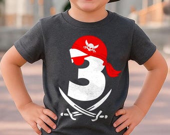 May include: A dark grey short-sleeved t-shirt featuring a large white number three graphic. The number is adorned with a red pirate bandana, a black eye patch, and two crossed white swords positioned underneath the number design.