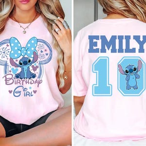 May include: Pink t-shirt featuring a blue cartoon character with a large bow, surrounded by a Mickey Mouse-shaped design. The front says "Birthday Girl." The back has the name "EMILY" and the number "10" with a matching cartoon character.