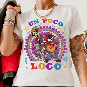 May include: White t-shirt featuring a colorful graphic of two skeleton characters from the movie Coco, with the words "UN POCO LOCO" in purple and blue. The design includes musical notes and floral accents.