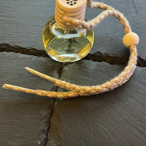 May include: A small, clear glass bottle with a wooden cap and a gold string. The cap has a logo of a stylized eagle with wings spread. The bottle is filled with a light yellow liquid.