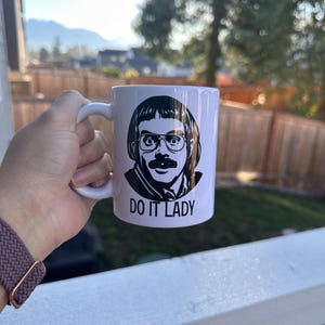 May include: White ceramic mug featuring a black and white illustration of a man with glasses and a mustache. The text "DO IT LADY" is printed below the illustration. The mug is held in a hand.