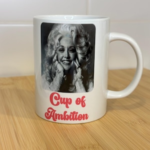 May include: White ceramic mug with a black and white photo of Dolly Parton. The mug has the text "Cup of Ambition" in pink.