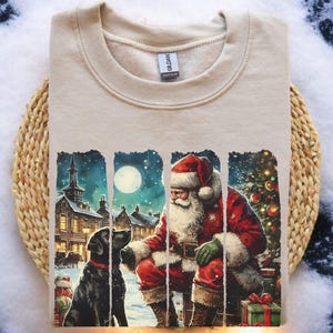 Puede incluir: Sudadera beige de cuello redondo con un gráfico navideño. El diseño representa a Papá Noel, un perro negro, un pueblo nevado y un árbol de Navidad. El diseño está enmarcado por paneles rectangulares verticales.