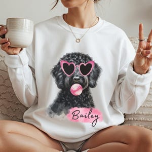 Può includere: Felpa bianca con un cane nero con occhiali da sole rosa a forma di cuore e una bolla rosa. Il nome "Bailey" è scritto in corsivo su uno sfondo rosa acquerello.