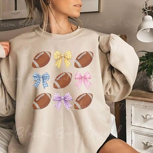 Puede incluir: Una sudadera beige con un dise&ntilde;o gr&aacute;fico de balones de f&uacute;tbol marrones y lazos de cuadros de colores.