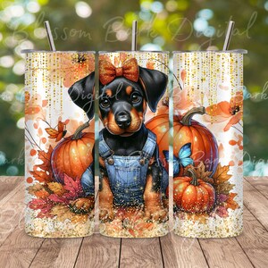May include: A black and brown Doberman Pinscher puppy wearing a blue denim overall dress with a red bow. The puppy is surrounded by fall leaves and pumpkins. The image is on a white background with gold glitter.