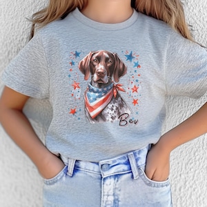 Puede incluir: Camiseta gris claro con una ilustración de un perro con un pañuelo patriótico. El perro es marrón y blanco con el nombre "Bev" debajo de la imagen. Estrellas rojas y azules rodean al perro.