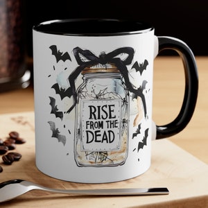 Peut inclure: Une tasse en céramique blanche avec une anse et un bord noirs. La tasse présente une illustration en noir et blanc d'un bocal avec un nœud noir et des chauves-souris. Le bocal porte le texte "Rise from the Dead".