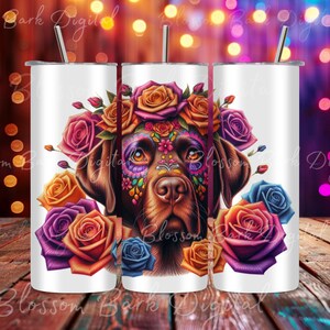 Pode incluir: Um copo branco de aço inoxidável com um design colorido inspirado no Dia dos Mortos. Mostra um cão castanho com maquiagem de caveira de açúcar e uma coroa de flores. Os olhos do cão são azuis e as flores são laranja, roxo e azul.