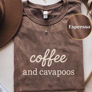 Könnte beinhalten: Espressofarbenes T-Shirt mit den Worten "coffee and cavapoos" in weißer, kursiver Schrift. Ein brauner Hut und eine silberne Halskette mit einem kleinen Anhänger sind ebenfalls zu sehen. Das Shirt liegt auf einer weißen Oberfläche.