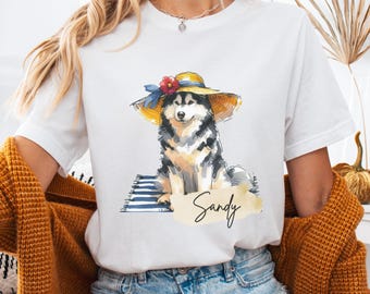Puede incluir: Camiseta blanca con una ilustración de acuarela de un perro con un sombrero de sol amarillo con una flor roja y una cinta azul. El perro está sentado en una toalla de playa a rayas. El nombre "Sandy" está escrito en cursiva.
