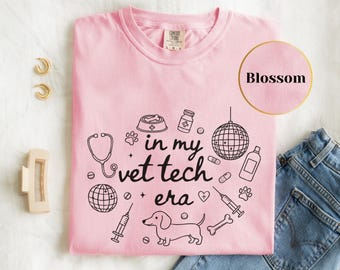 May include: A light pink t-shirt with the phrase "in my vet tech era" and various veterinary-themed graphics in black. The graphics include a stethoscope, dog, pills, and a disco ball. The shirt is a Comfort Colors brand.
