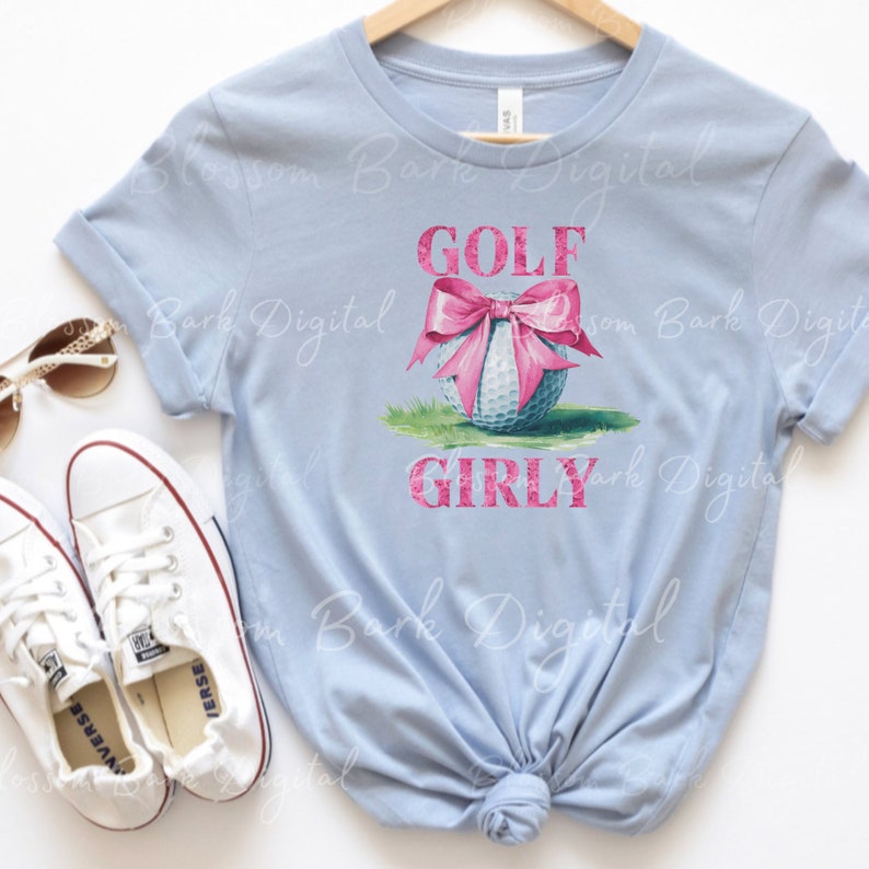 K&ouml;nnte beinhalten: Hellblaues T-Shirt mit einem wei&szlig;en Golfball, auf dem ein rosa Schleife sitzt. Der Text "GOLF GIRLY" ist in rosa Glitzer auf das Shirt gedruckt.
