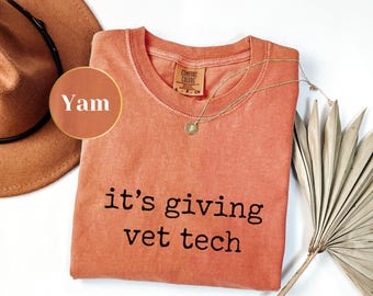 May include: A coral-colored t-shirt with the phrase "it's giving vet tech" printed in black. A gold necklace with a pendant rests on the shirt. A brown hat and a dried palm leaf are also visible.