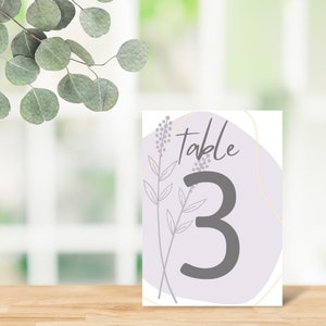 Pode incluir: Um cartão de número de mesa branco com um design abstrato roxo e dourado. O cartão diz "Mesa 3" em texto preto.