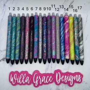 May include: A collection of seventeen glitter pens in various colors, including teal, purple, pink, and silver. Each pen has a unique design and a black tip. The image also features a pink sticker with the text "Willa Grace Designs."