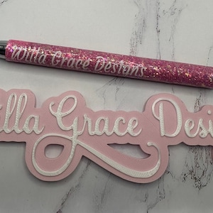 May include: Pink glitter pen with the text "Willa Grace Designs" on it, next to a pink sign with the same text in white raised lettering.